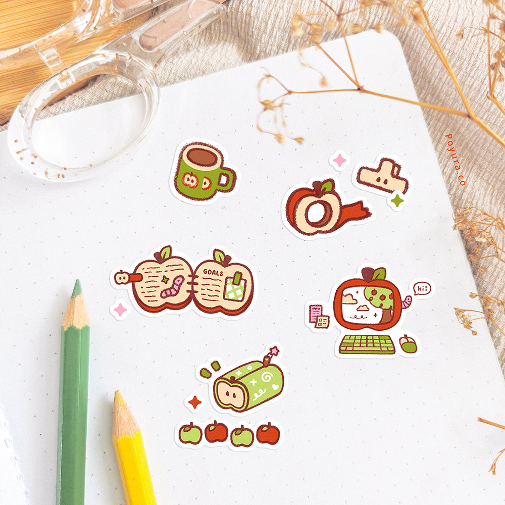 A notebook is decorated with apple stationery-themed stickers. The desk is decorated with pencils, a scissor, and some dried flowers.