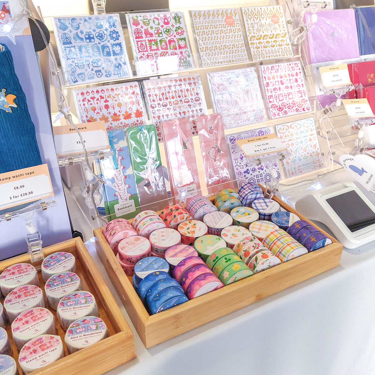 A table displaying stationery items. There are washi tapes displayed in a wooden tray. Behind it is a display for sticker sheets and bookmarks.