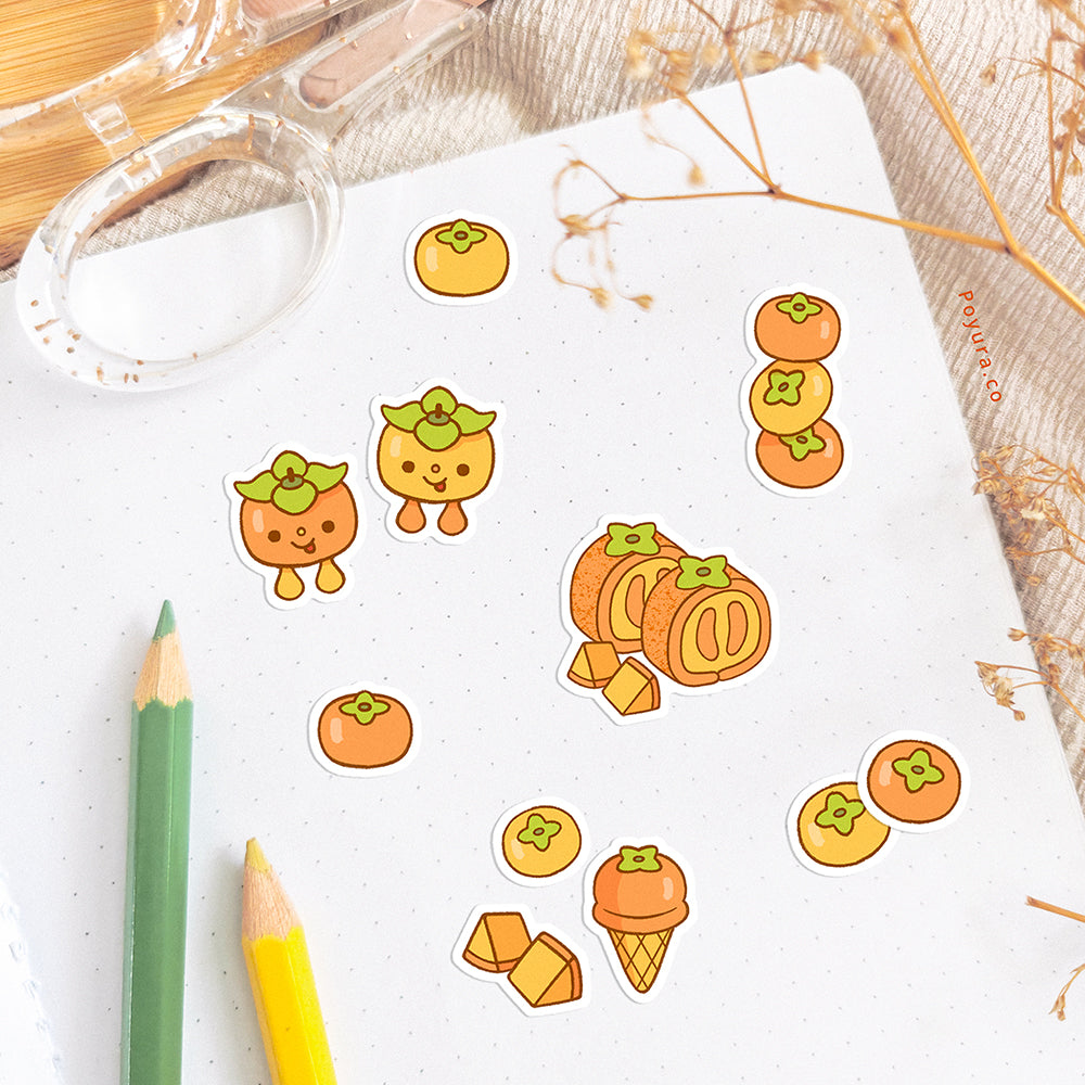 A notebook is decorated with persimmon stickers. The desk is decorated with pencils, a scissor, and some dried flowers.