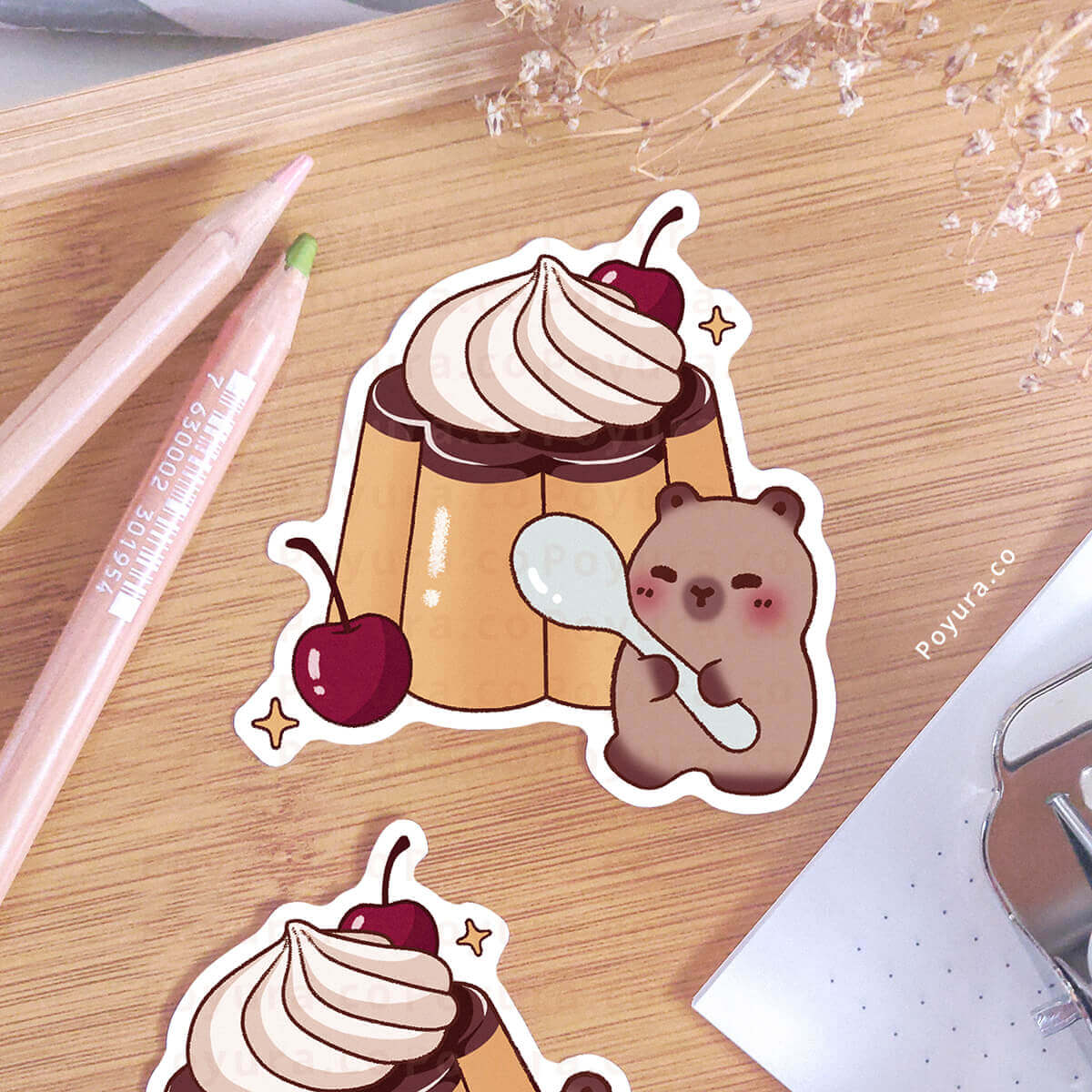 Sticker of a pudding with a cherry and a capybara holding a spoon on a wooden surface.