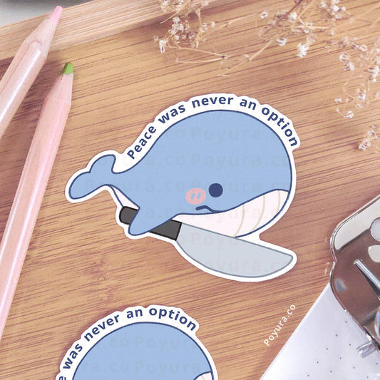 Sticker of a whale with a knife and 'Peace was never an option' text on a wooden surface with a pen and notebook.